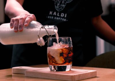 A barista skillfully pours milk into a glass of iced coffee on a wooden board.